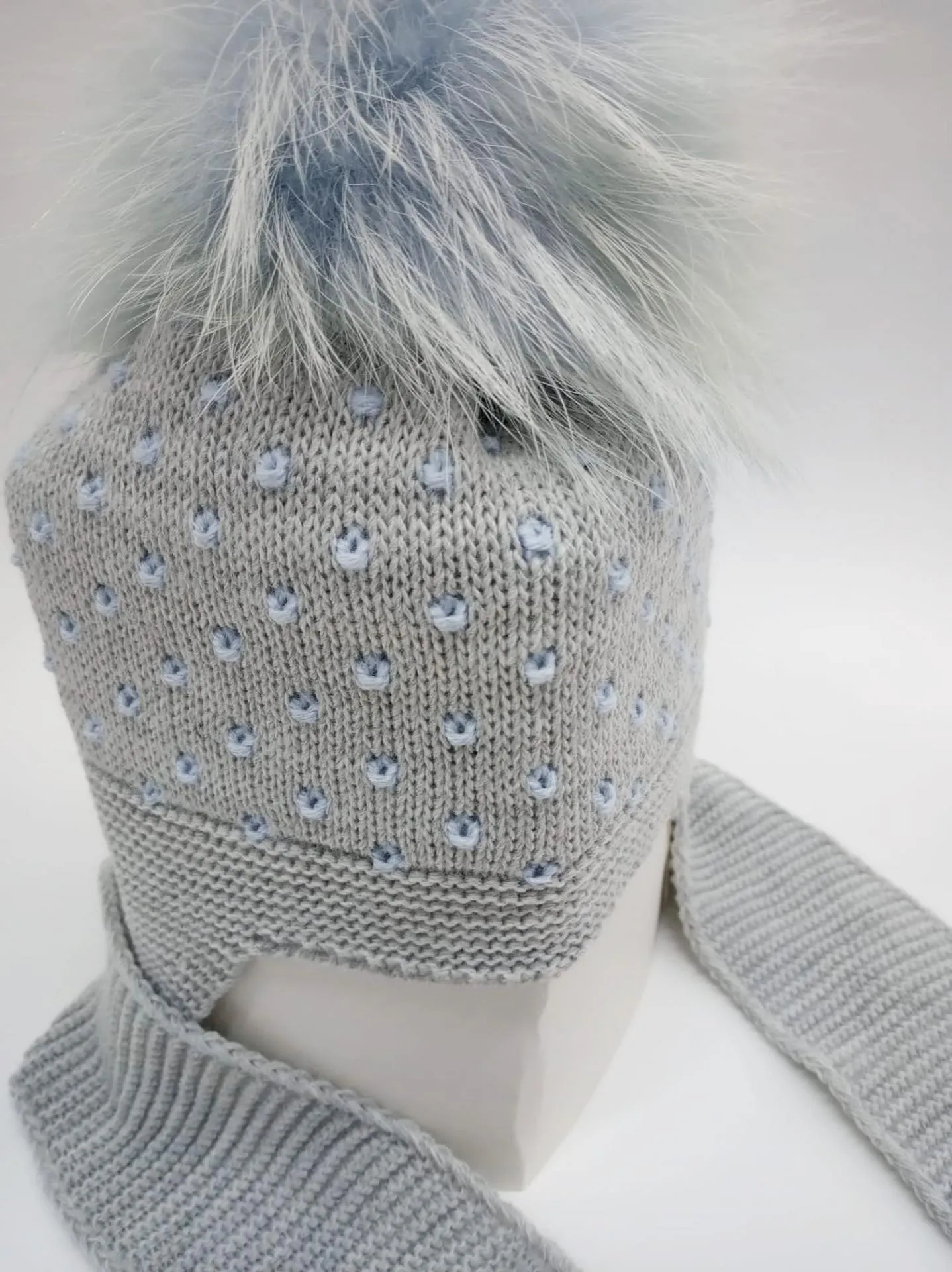 Gorro gris con motas azules con bufanda unida y pompóm de pelo natural.